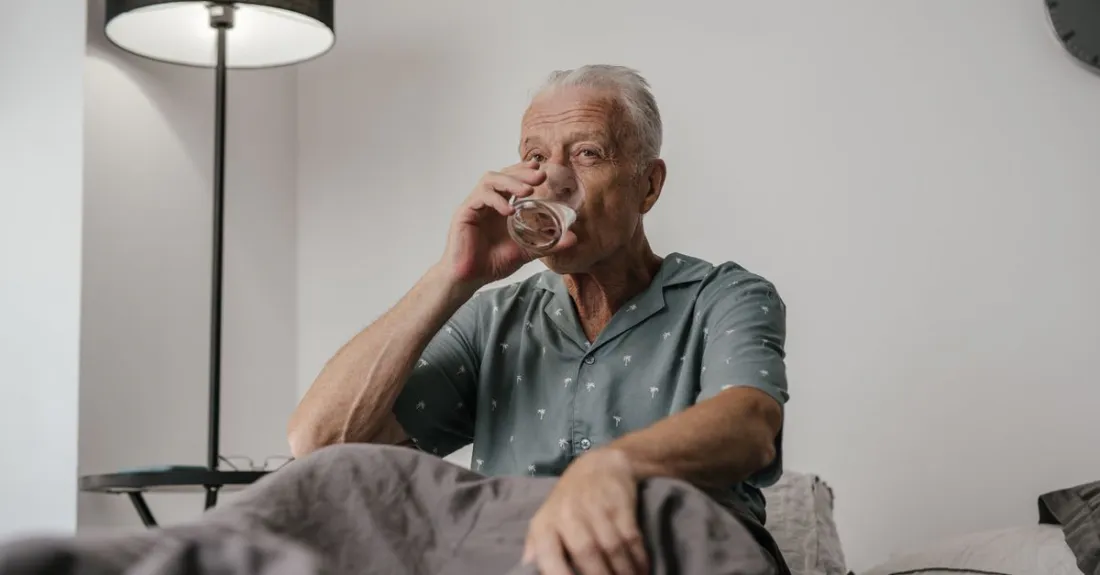 Older man sitting up in bed at night drinking a glass of water